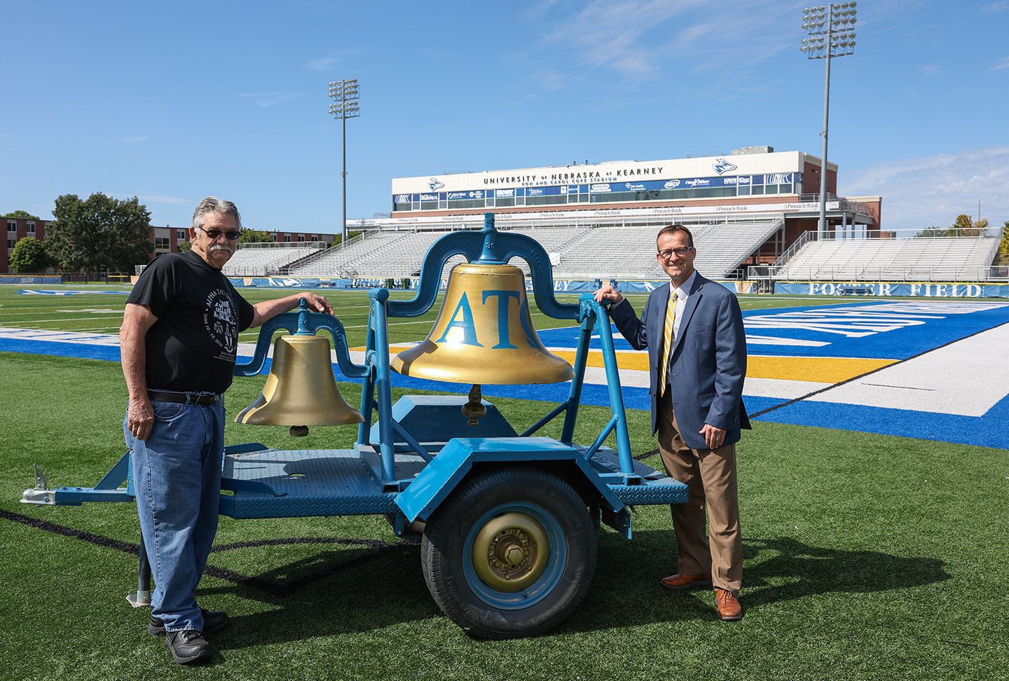 Former Phi Tau Gamma/Alpha Tau Omega fraternity member Tom Paxson, left, and UNK Athletic Director Marc Bauer are pictured with the Loper Victory Bell at Cope Stadium. (Photo by Erika Pritchard, UNK Communications)