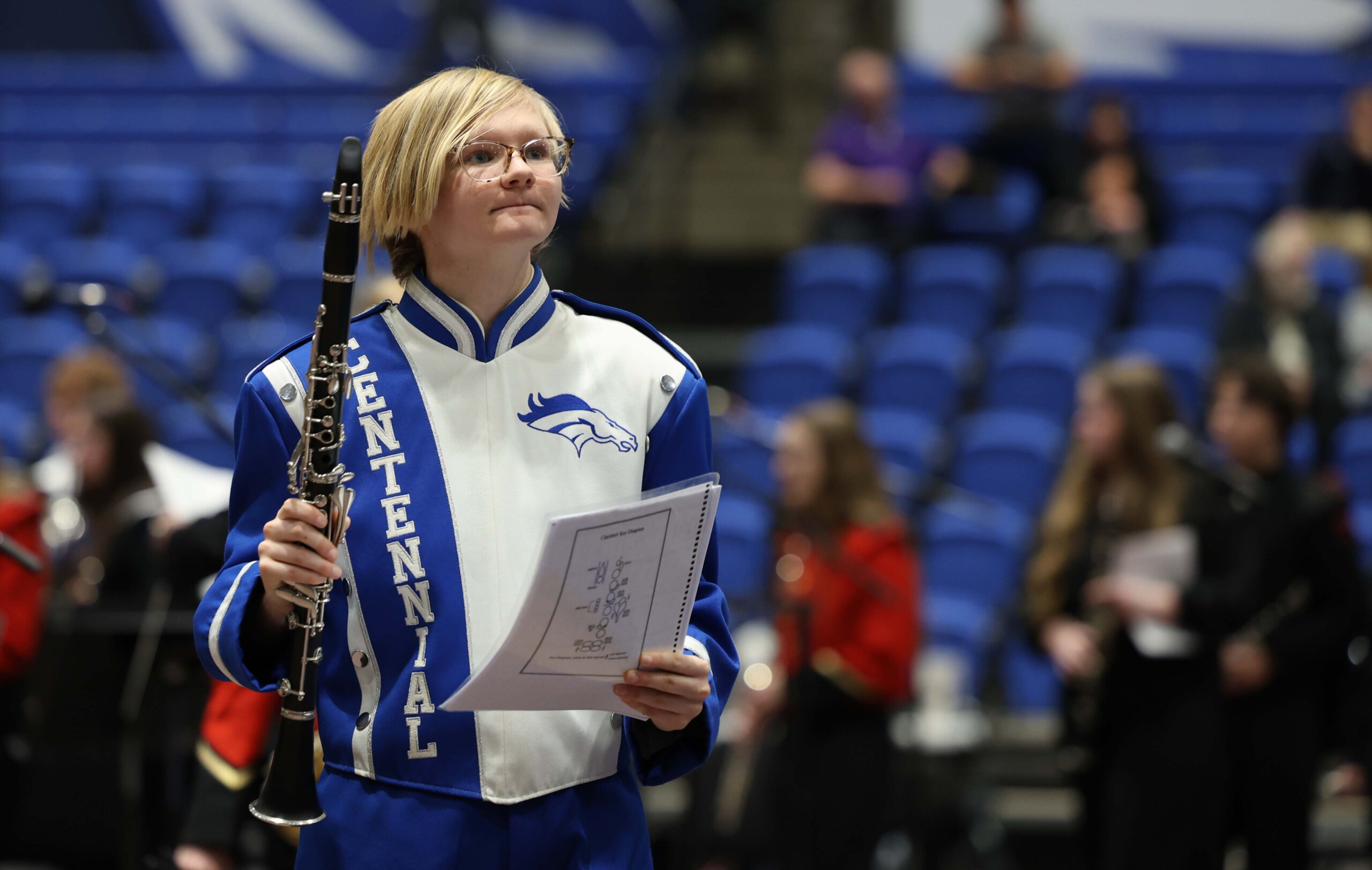 A high school student in a band uniform walks with her clarinet and music sheets.