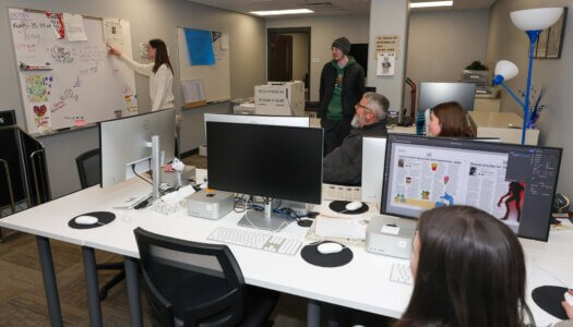 A student writes on a whiteboard while others look on in the newspaper room.