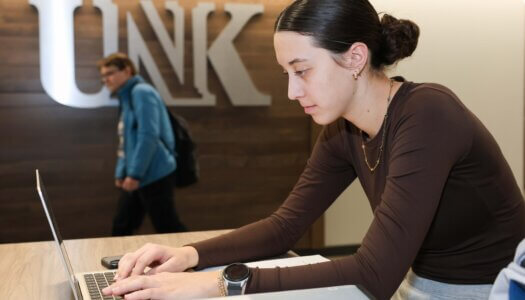 A student works on her laptop while another walks by in the library.