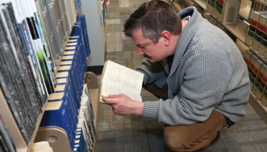A professor reads a book in an aisle at the library.