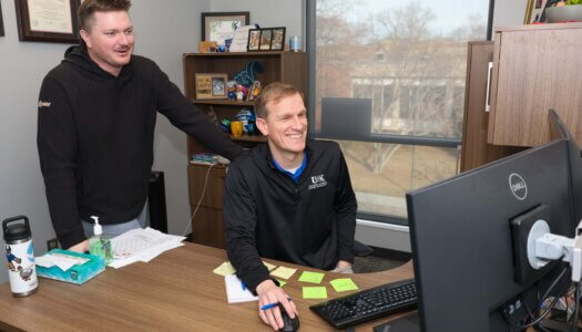 Two men look at a computer in an office.