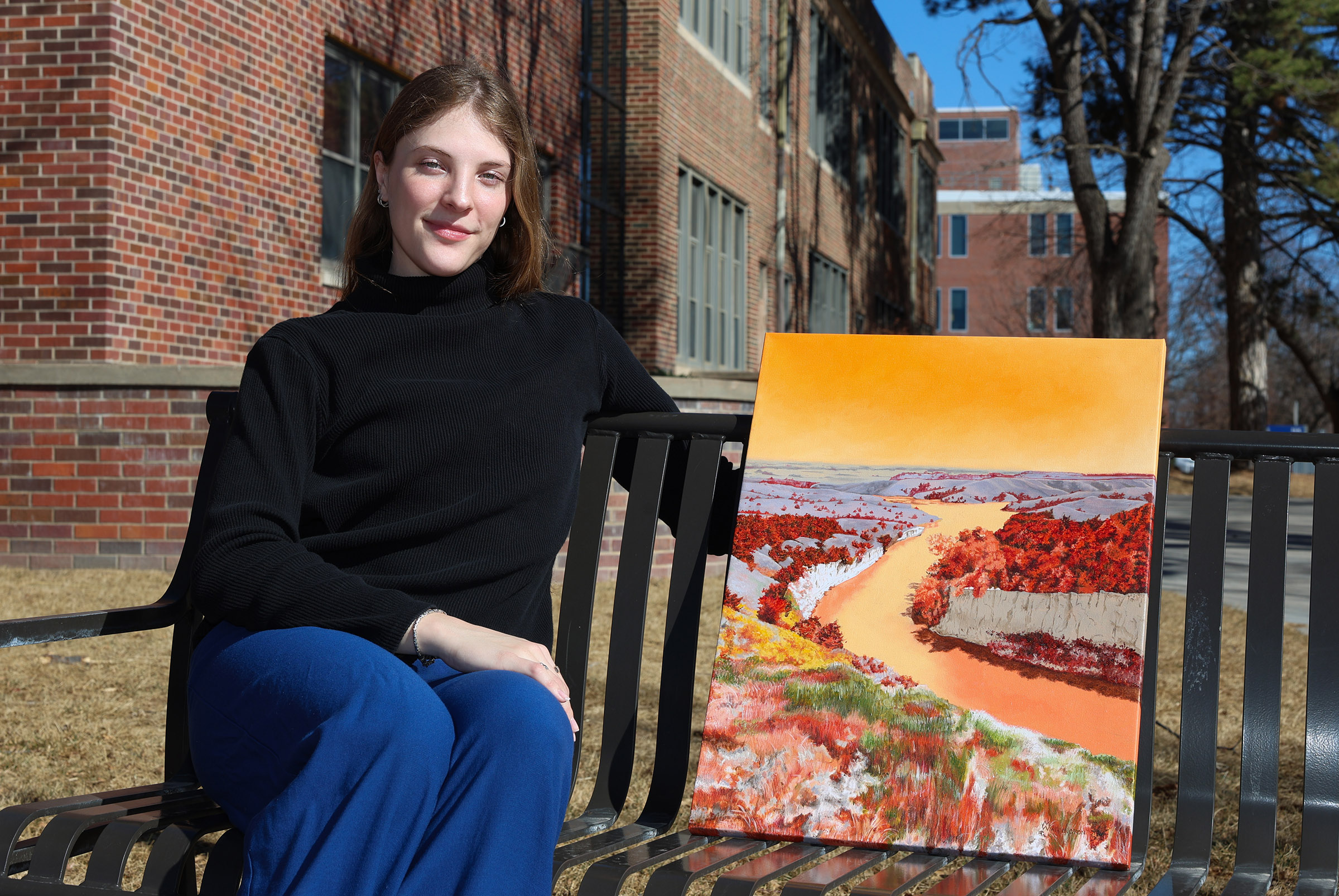 Maggie Waddington is pictured seated on a bench on campus, next to one of her paintings.