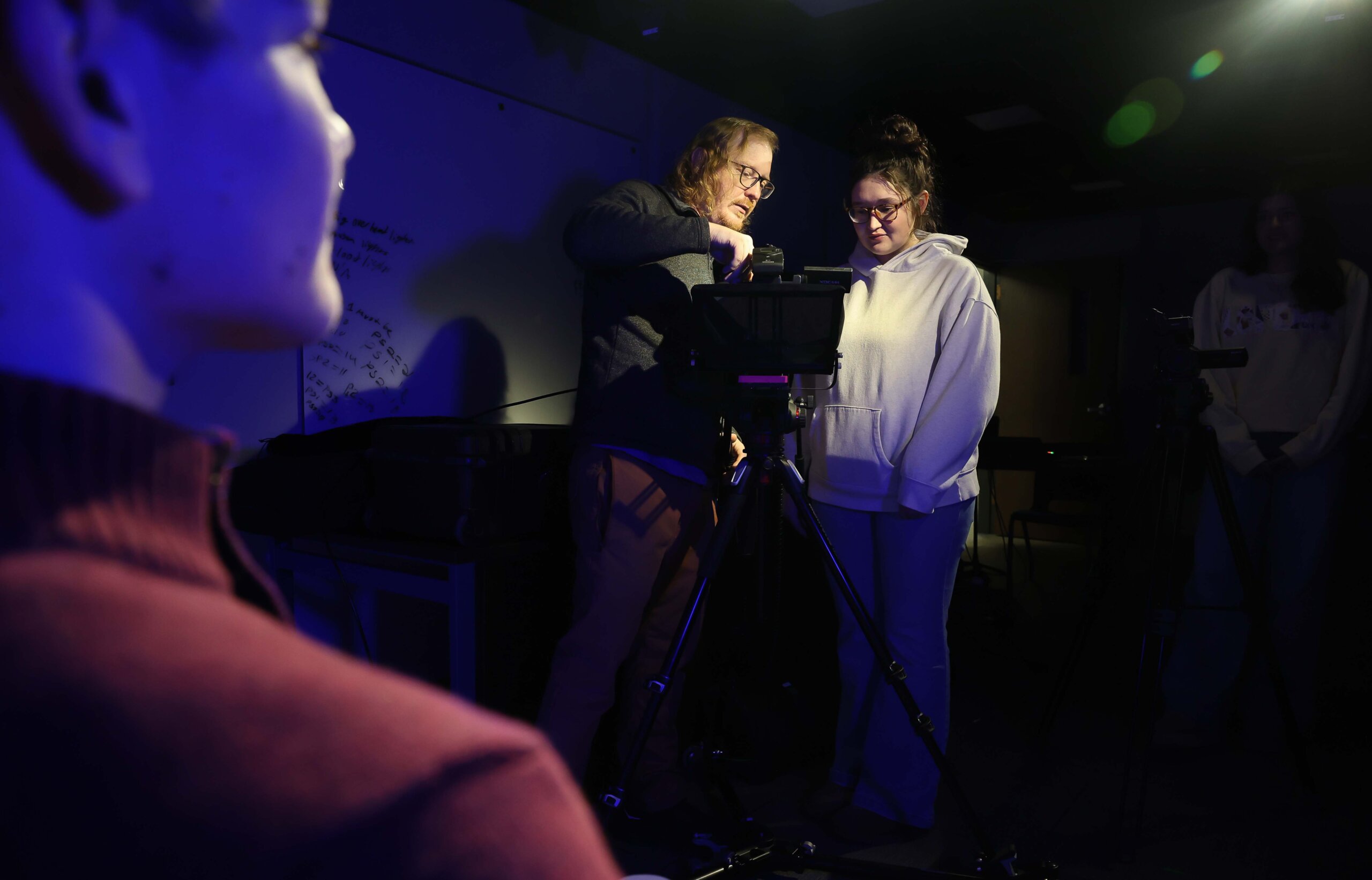 A professor shows a student how to operate a video camera while the video subject waits.