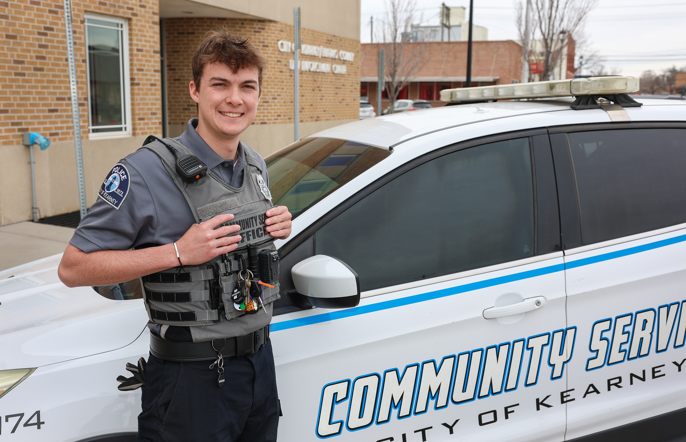 Wearing his community service officer uniform, Zach Arner poses for a photo with his vehicle outside the law enforcement center.