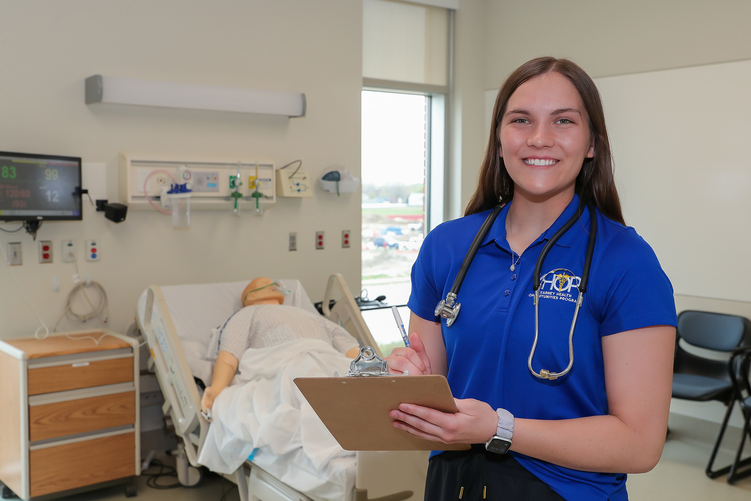 A KHOP student poses for a photo in the nursing lab, holding a clipboard.