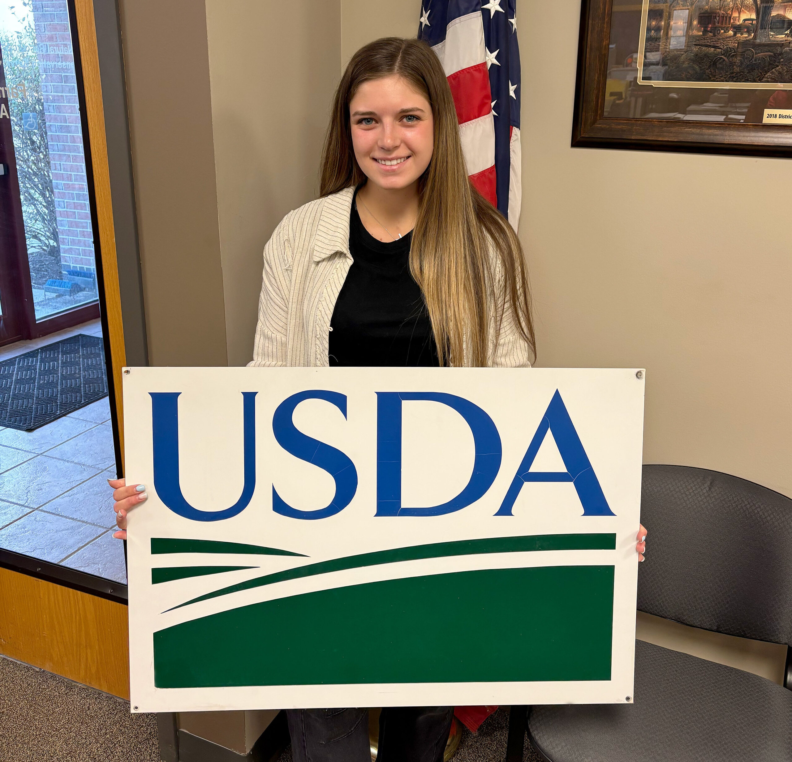 Karlie Wies poses for a photo at the Farm Service Agency, holding a USDA sign.