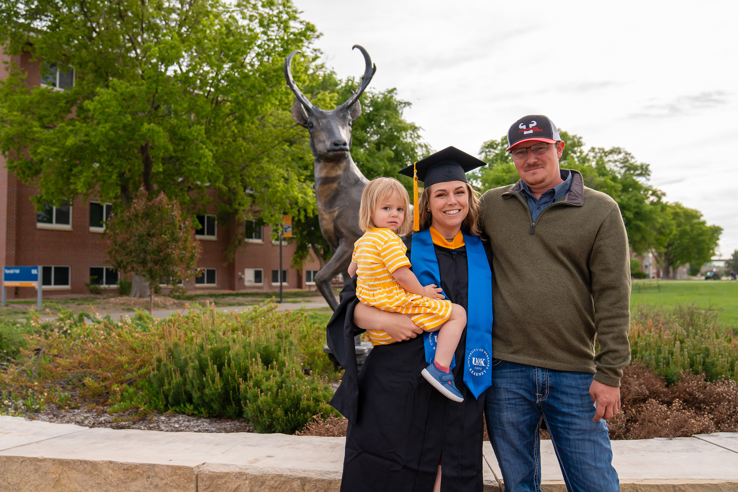 Wearing her graduation cap and gown, Leighlynn Towne poses for a photo with her husband and daughter on the UNK campus.