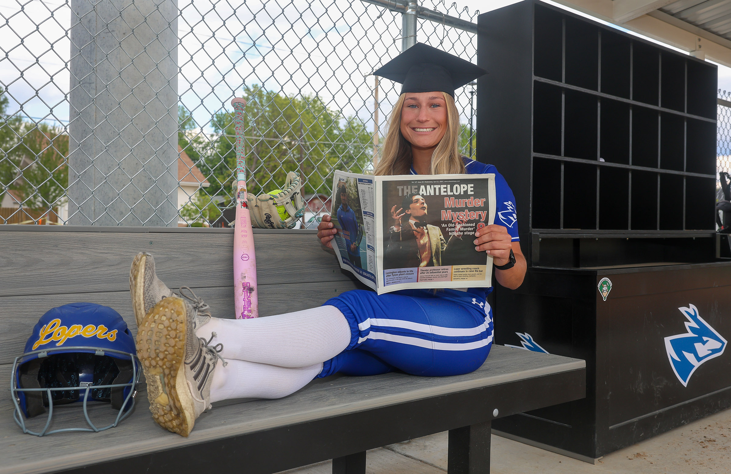 Mary Chvatal poses for a photo in the dugout, holding a newspaper and wearing her graduation cap.