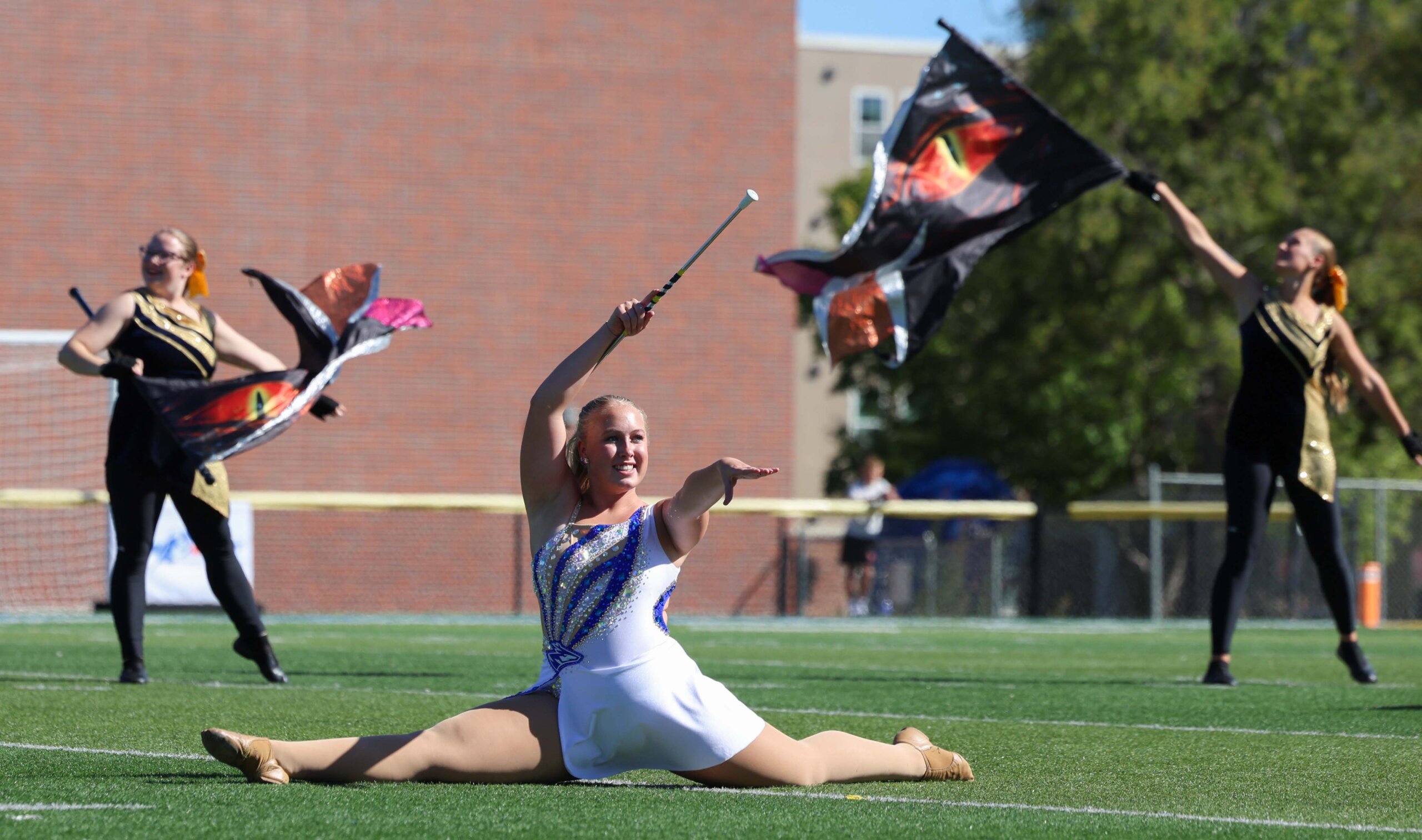 A baton twirler does the splits while girls twirl flags around her.